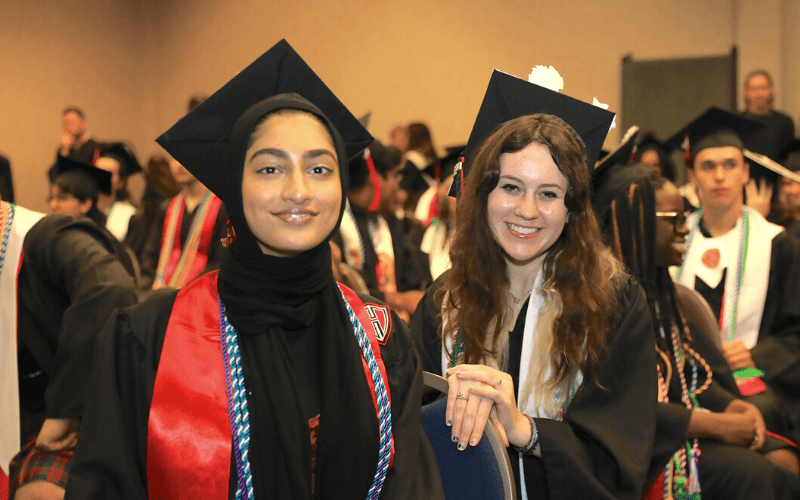 Two female students dressed in graduation robes and mortarboards smile to the camera